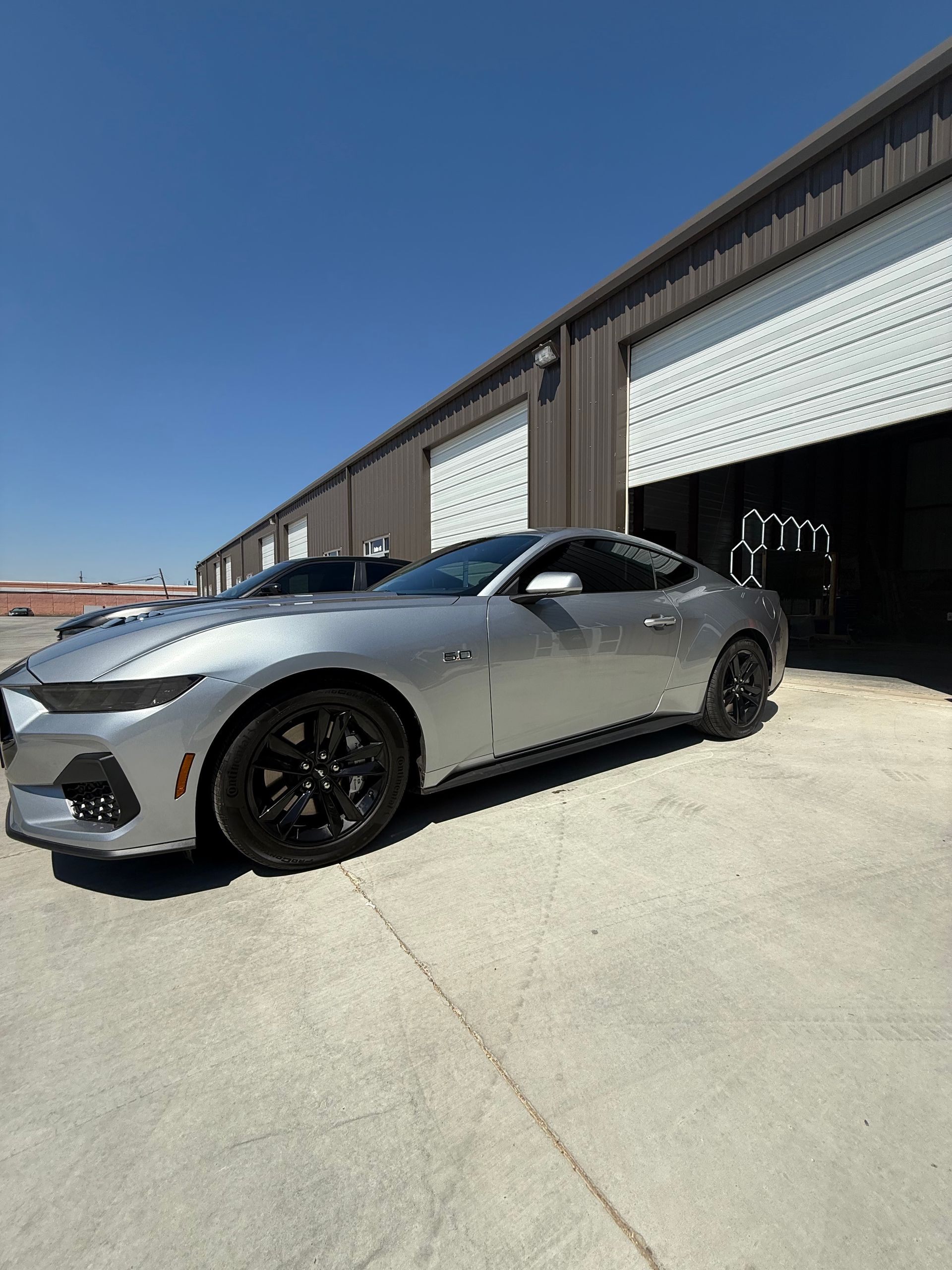 Silver Ford Mustang parked in front of a metal building with garage doors, under a clear blue sky.