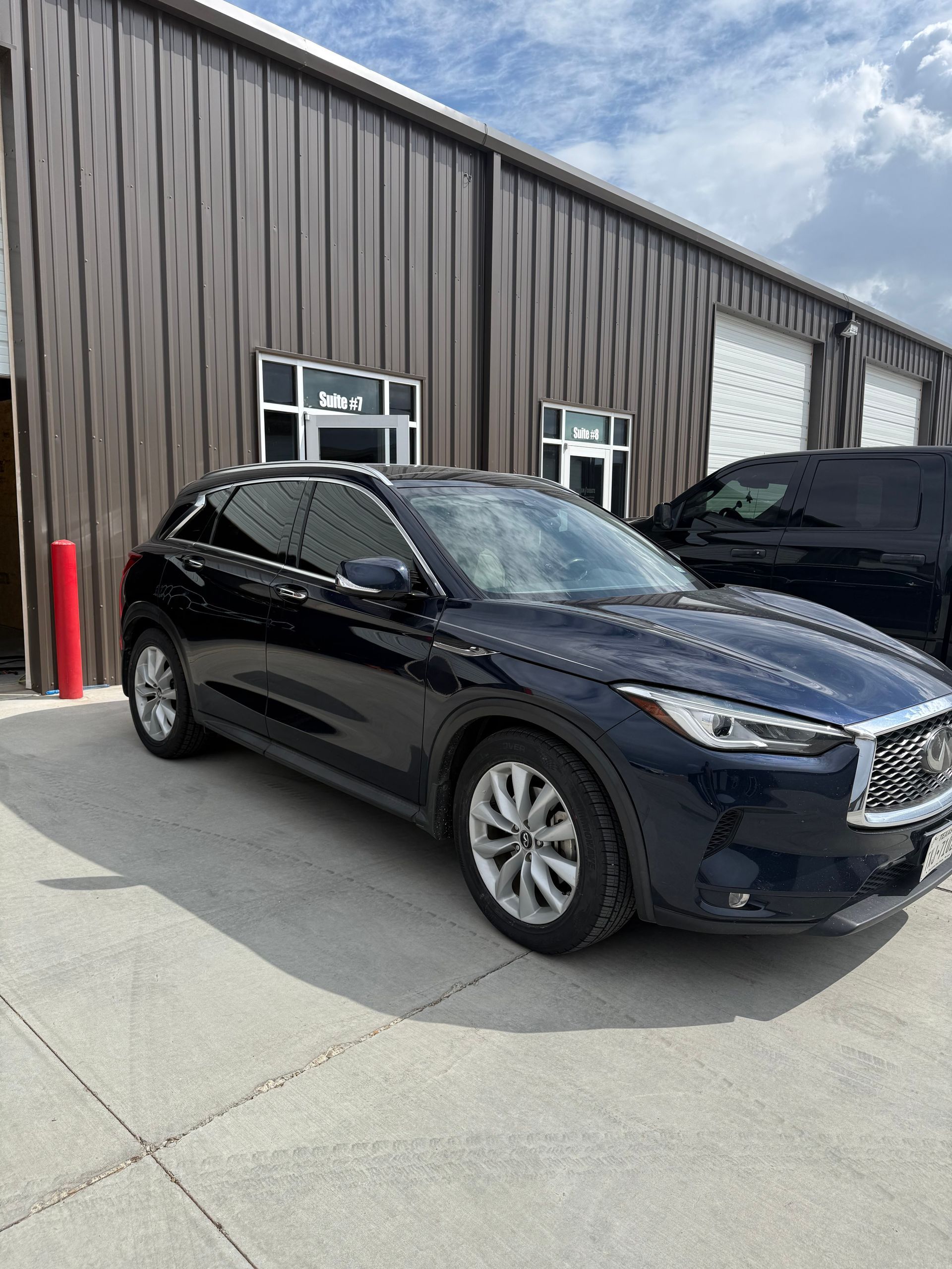 Dark blue Infiniti SUV parked in front of a brown building with windows; sunny day.
