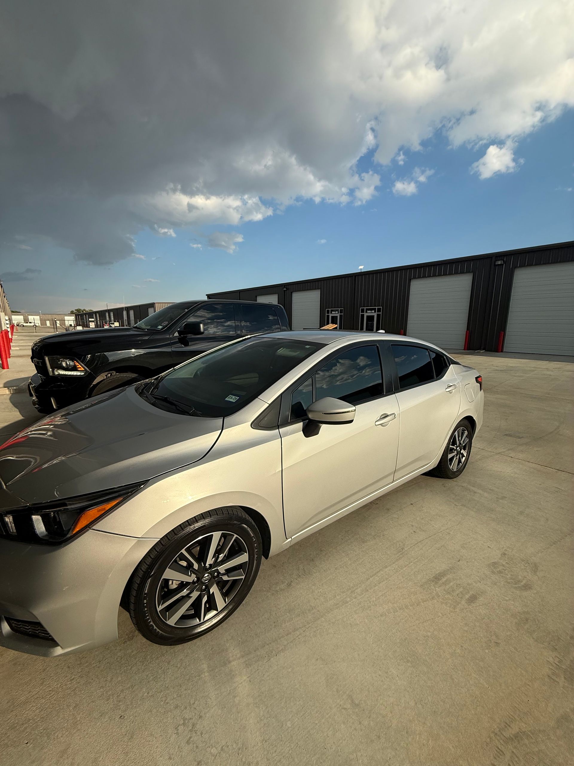 Silver car parked outside, with a black truck visible in the background against a backdrop of storage units under a cloudy sky.