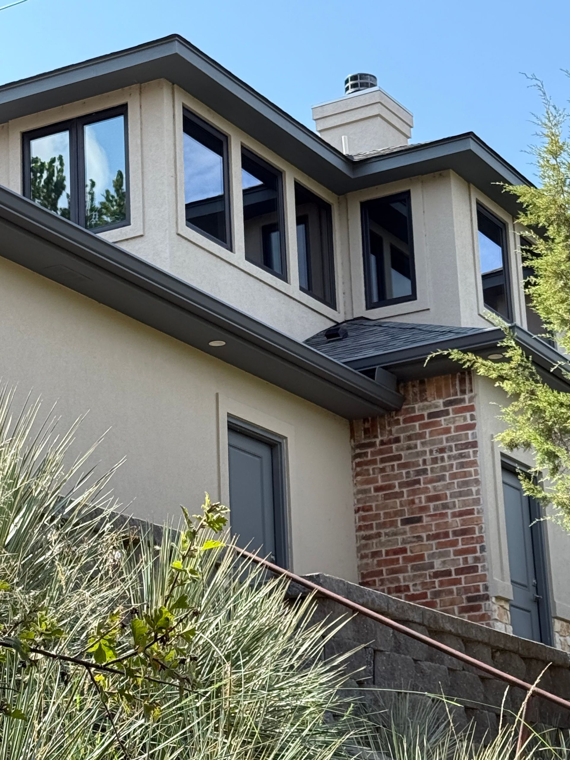 Two-story house with tan stucco, dark windows, and brick accents. Chimney on the roof.