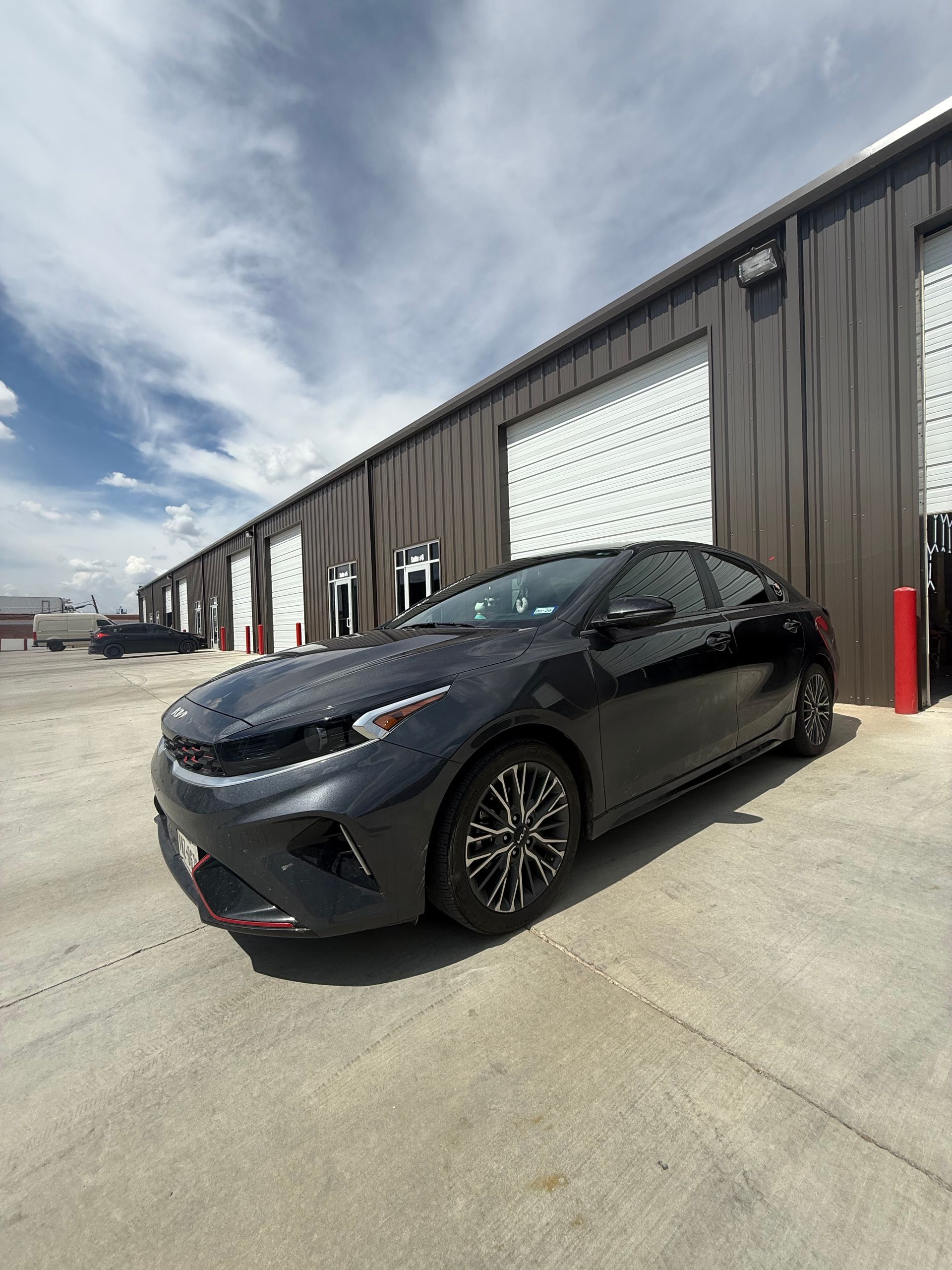 Dark gray Kia Forte parked in front of a building with roll-up doors, under a blue sky.