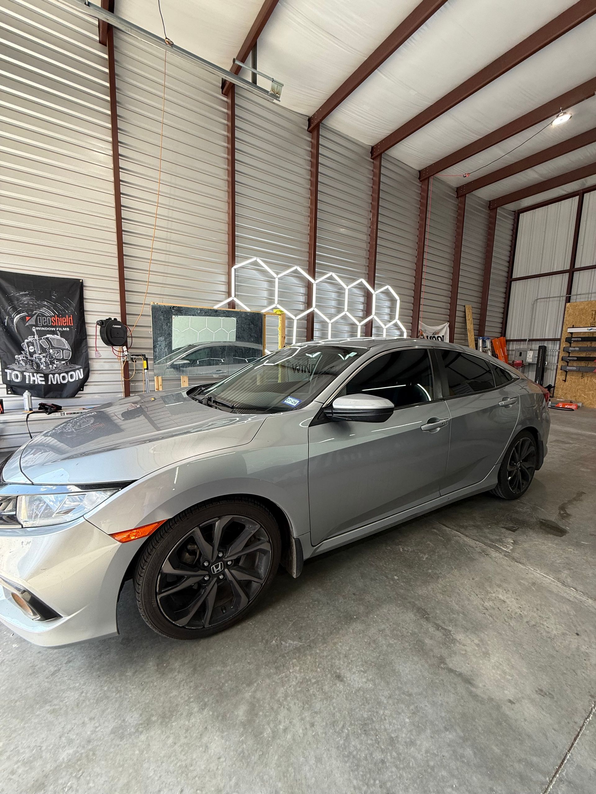 Gray Honda Civic parked inside a workshop. Black wheels, tinted windows. Hexagon lights on the wall.