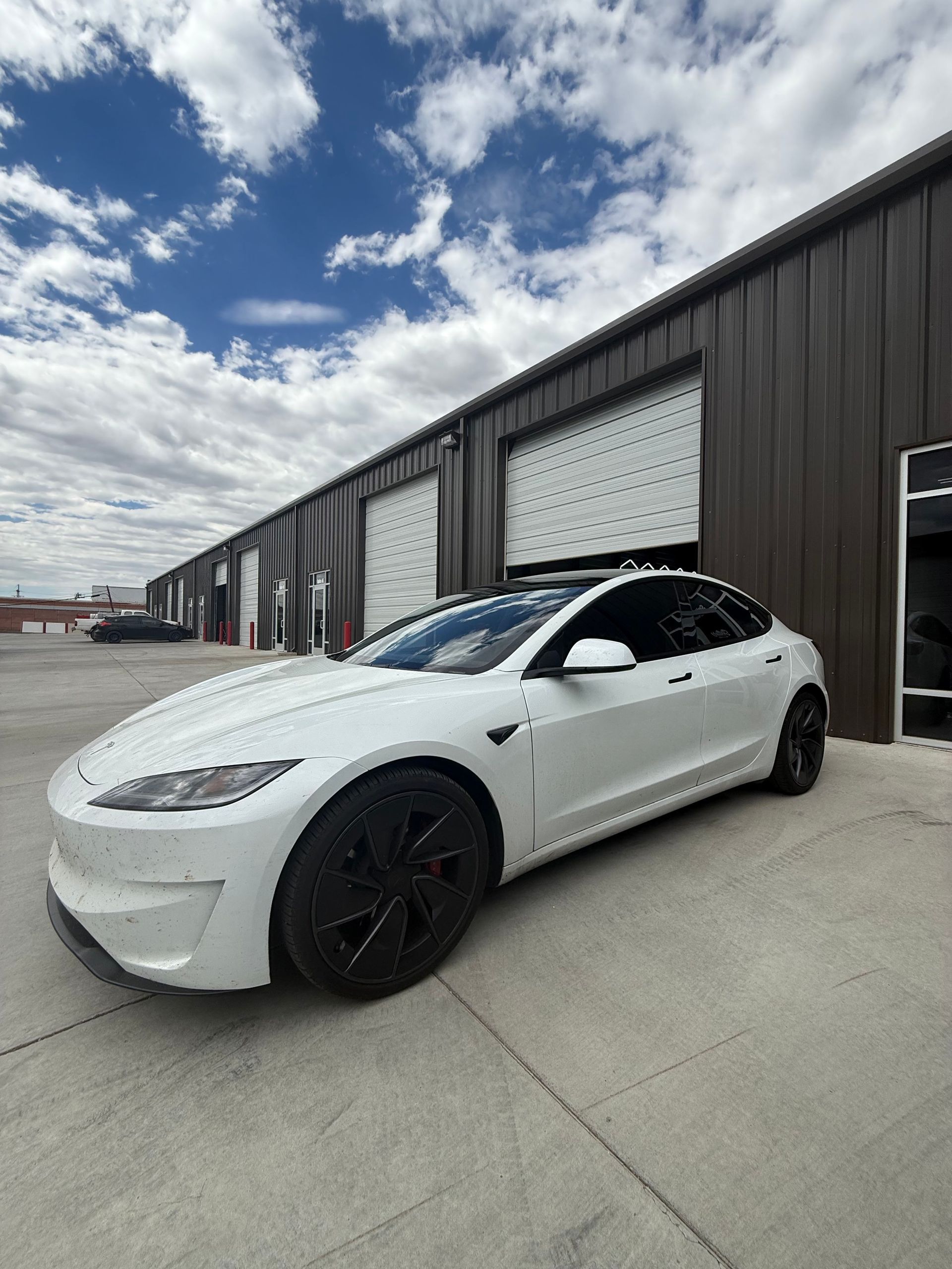 White Tesla parked in front of a building with roll-up doors, blue sky with clouds in background.