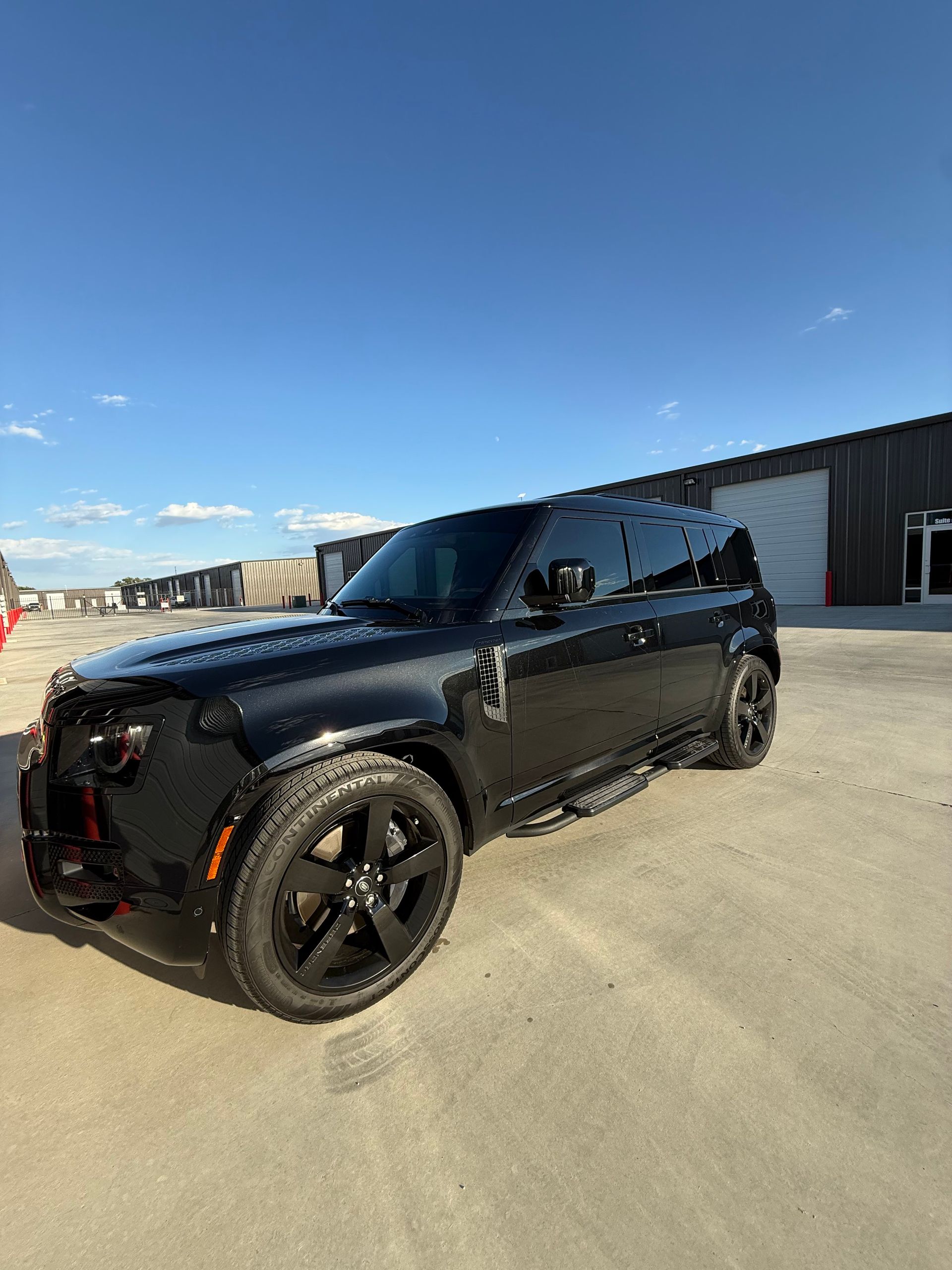 Black Land Rover Defender parked outdoors on concrete, against a blue sky.