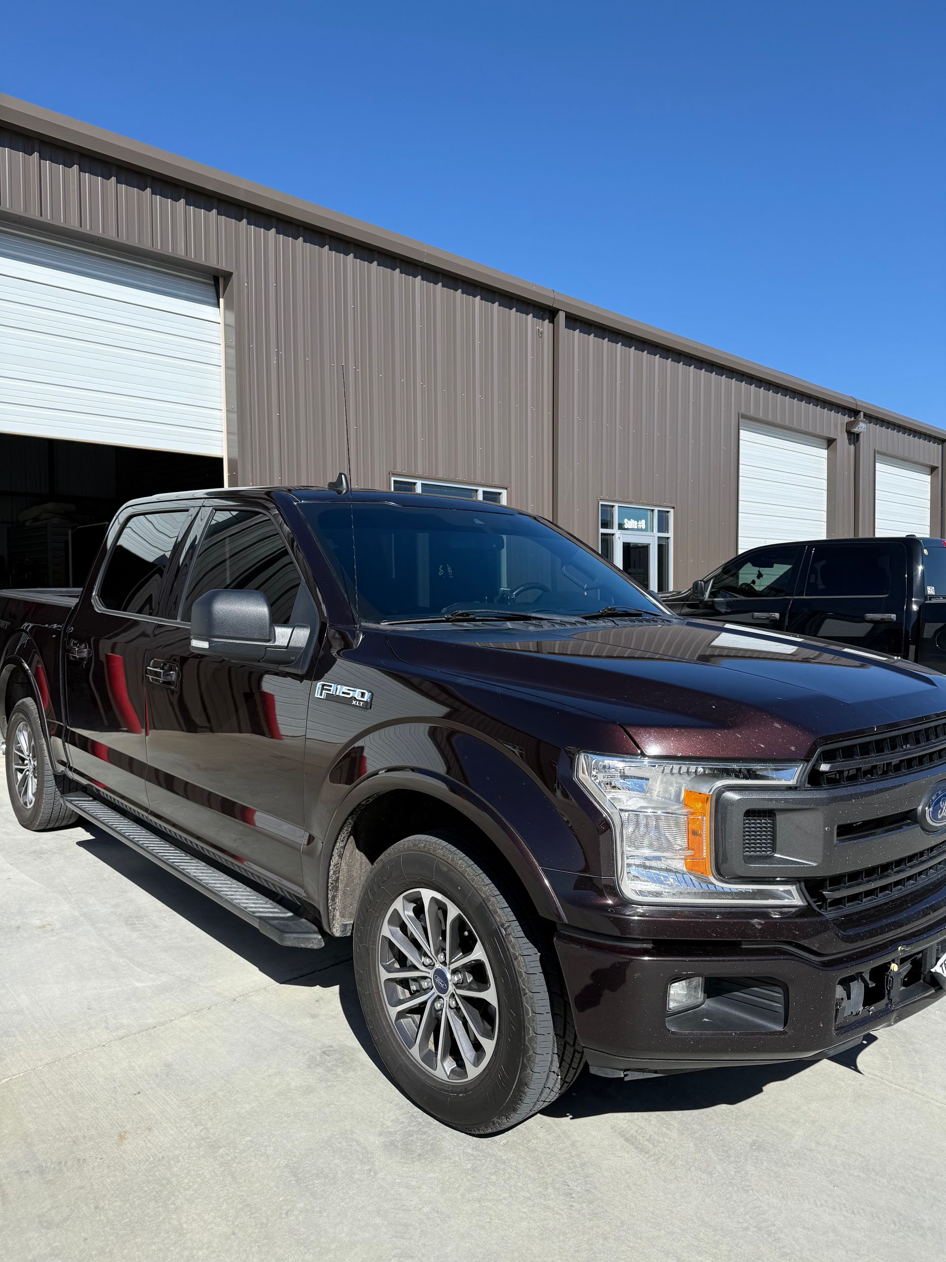 Dark brown Ford F-150 pickup truck parked in front of a brown building with a blue sky in the background.