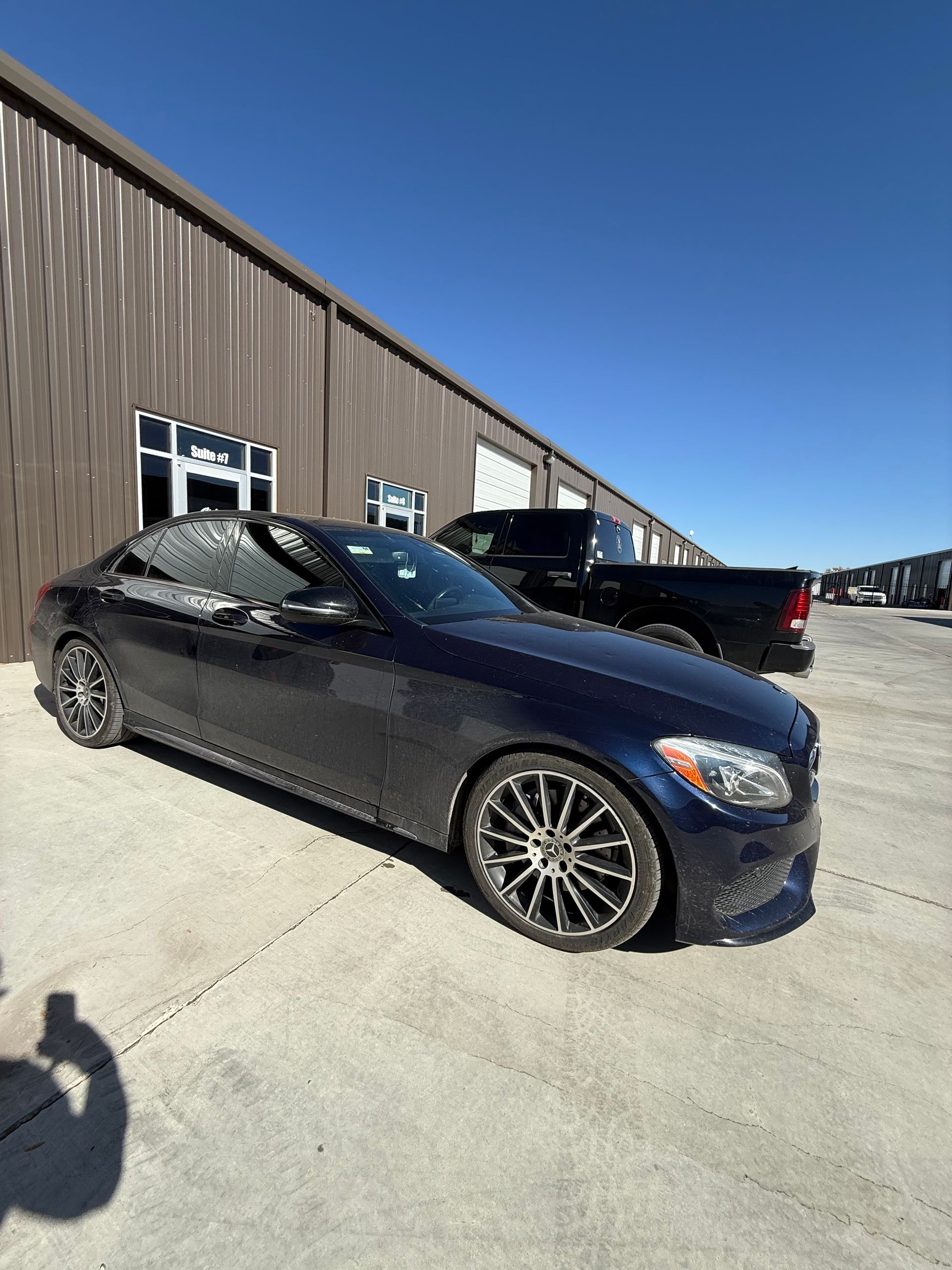 Dark blue Mercedes-Benz parked outside a metal building on a sunny day. A black truck is visible behind.
