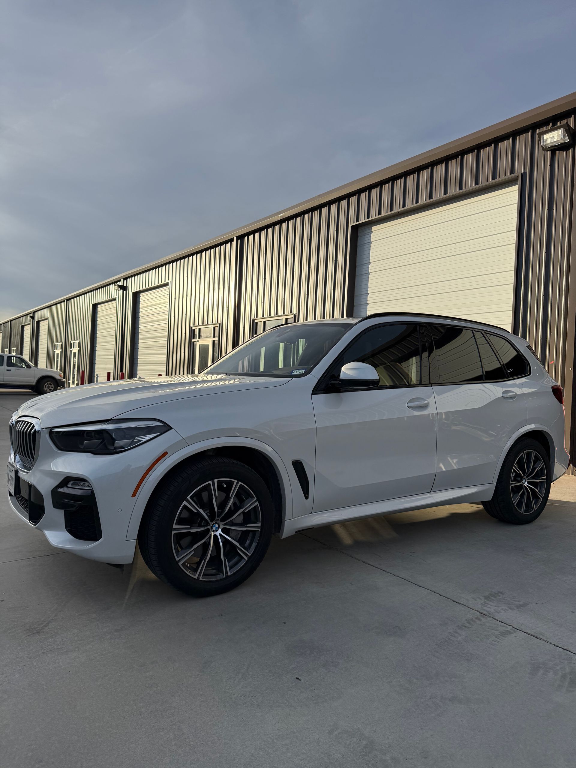 White BMW X5 SUV parked outside a building with garage doors. Black rims, overcast sky.