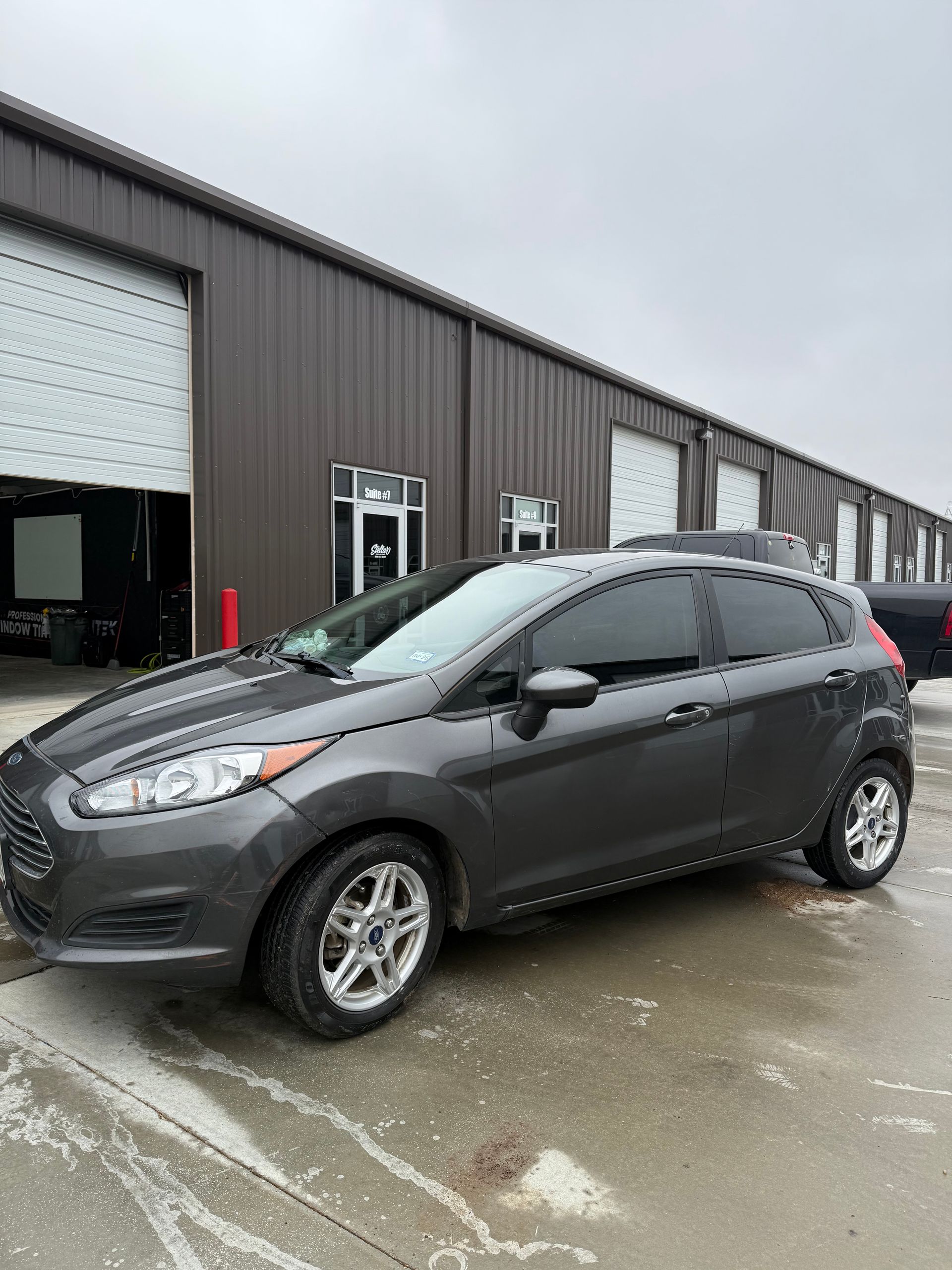 Gray Ford Fiesta parked in front of a brown building with garage doors. Overcast sky.