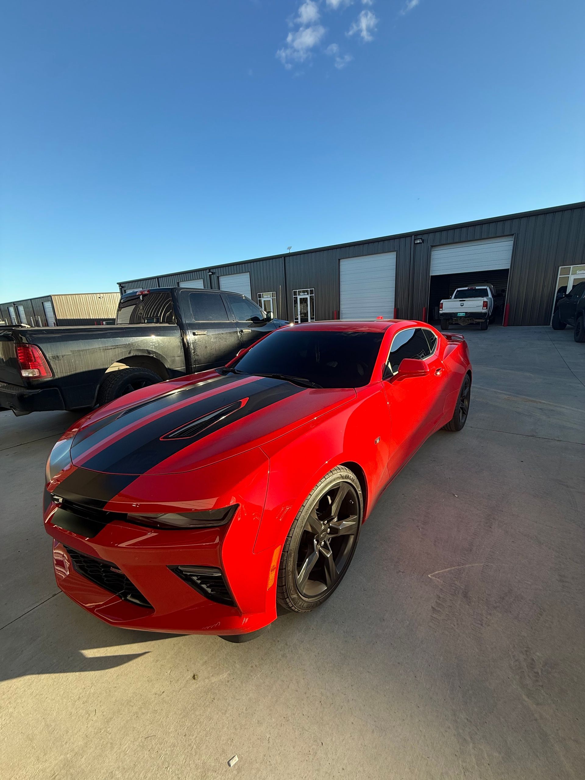 Red Chevy Camaro with black stripes parked outdoors, near a building and a black truck on a sunny day.
