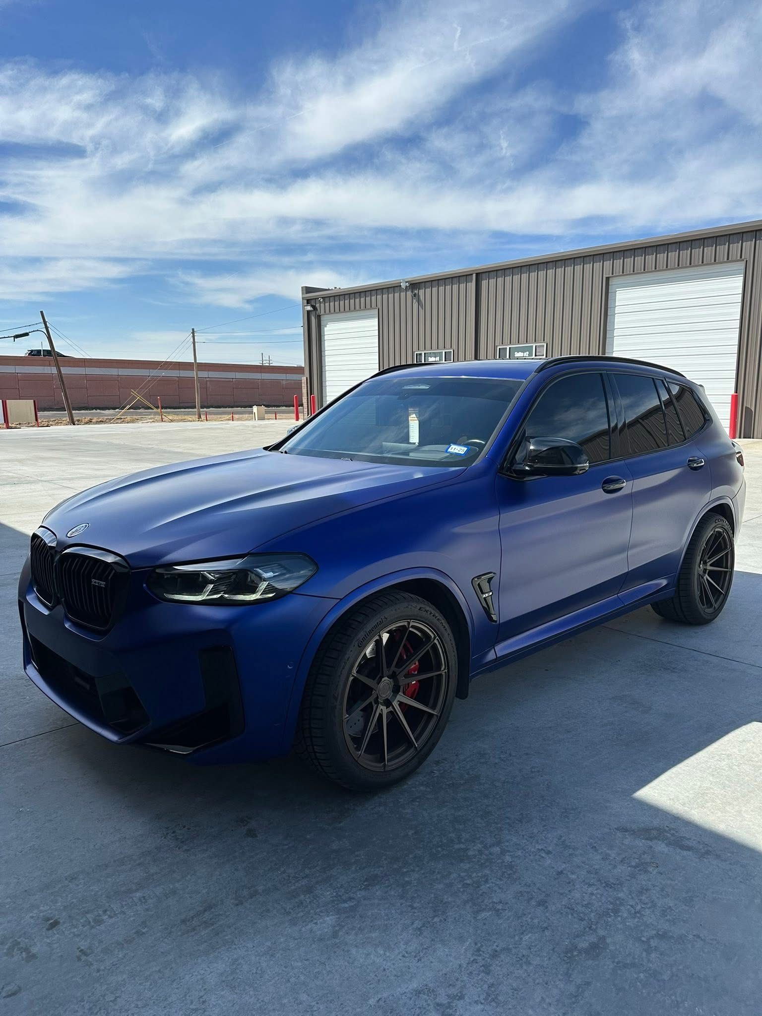 Blue BMW SUV with black accents parked on concrete; sunny day, industrial setting.