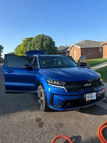 Blue Kia Sorento SUV with doors open, parked on a paved street in front of houses on a sunny day.