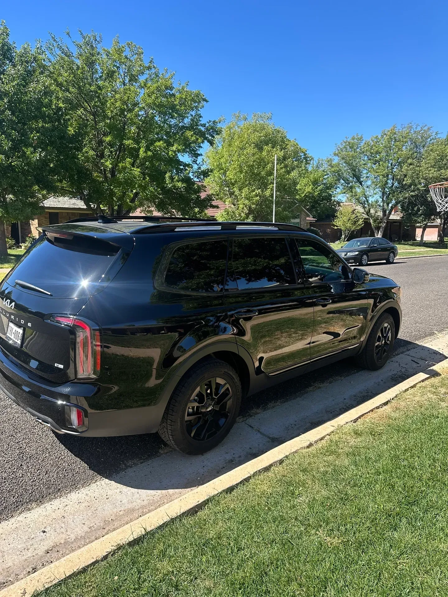 Black Kia SUV parked on a residential street next to green grass and trees under a blue sky.