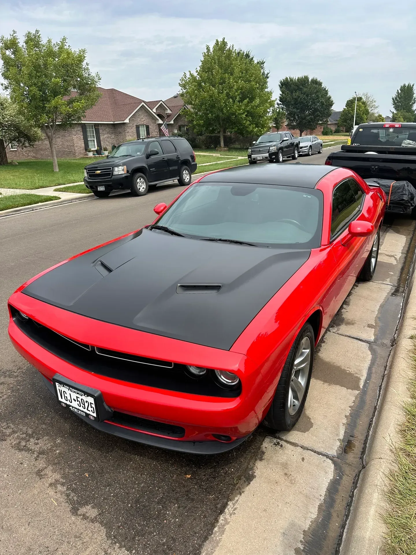 Red Dodge Challenger with a black hood parked on a street curb. Other vehicles in the background.