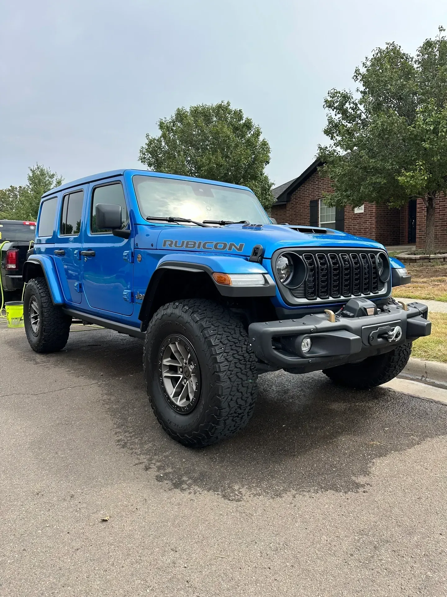 Blue Jeep Wrangler Rubicon parked on a paved street. Black grill and off-road tires. Cloudy sky.
