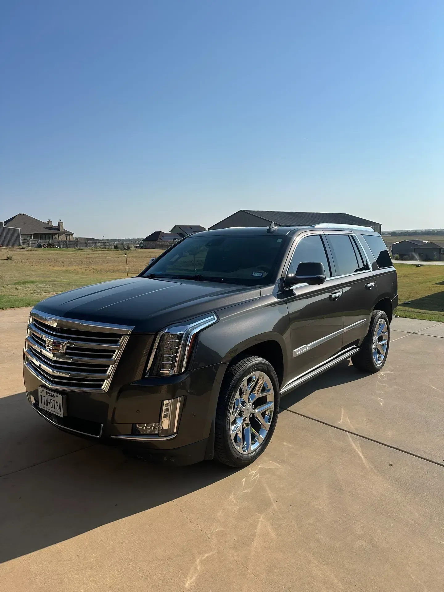 Dark gray Cadillac Escalade SUV parked on a concrete driveway with a clear blue sky in the background.