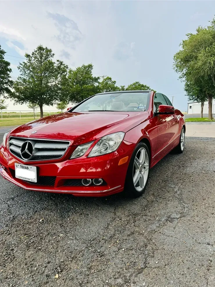 Red Mercedes-Benz convertible parked on gravel. Cloudy sky, trees in the background.