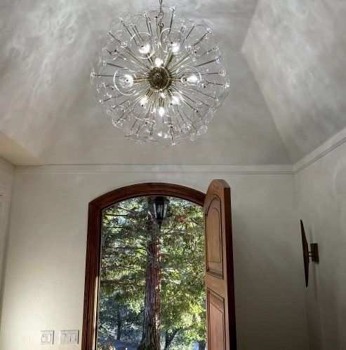 Ornate chandelier hangs above an open wooden door leading to a lush, green outdoor scene.