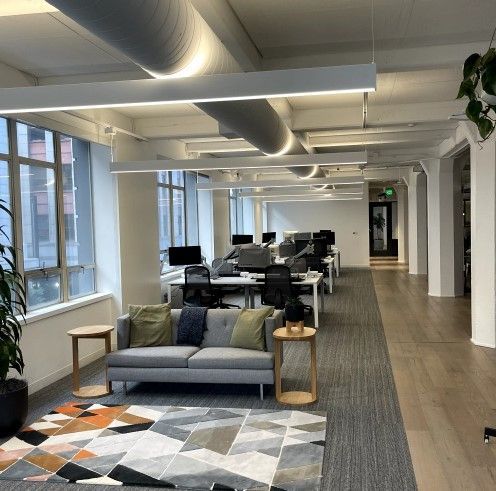 Modern office space with desks, sofa, rug, plants, and natural light from tall windows.