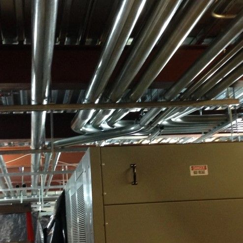 Electrical conduits and wires run above a metal electrical cabinet, in an unfinished ceiling.