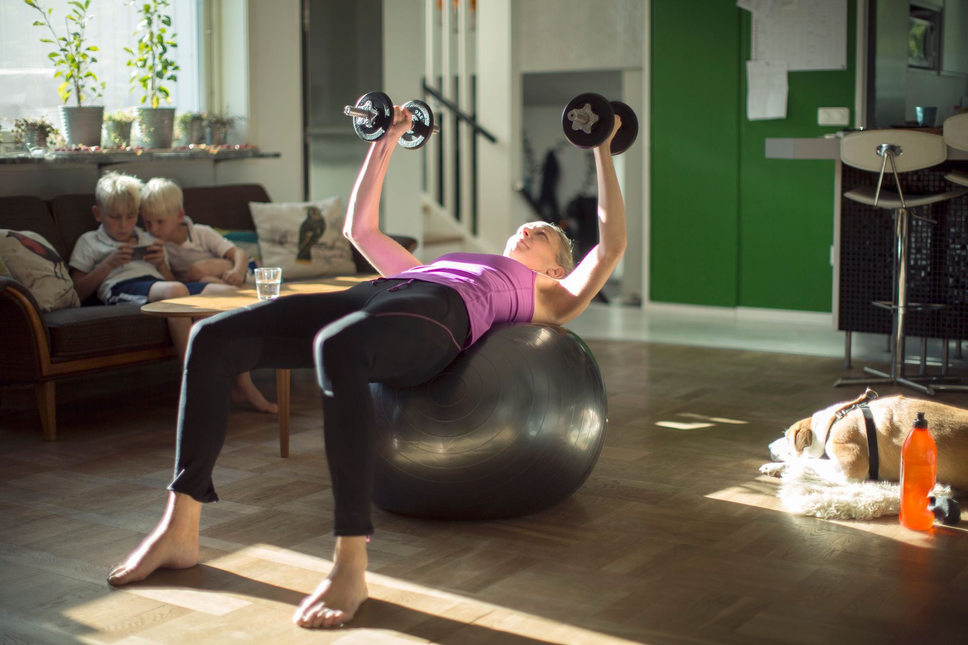A woman is lifting dumbbells on a pilates ball in a living room.