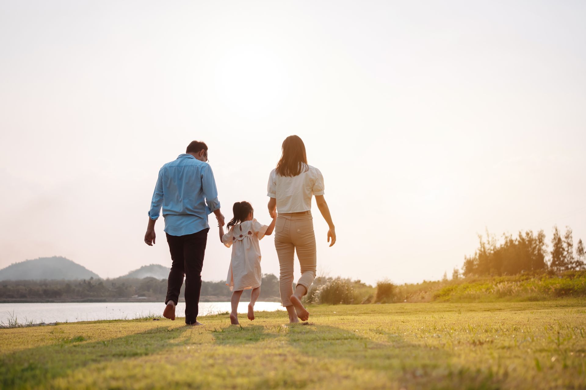 Family of three walking on grass near water, holding hands, silhouetted by the setting sun.