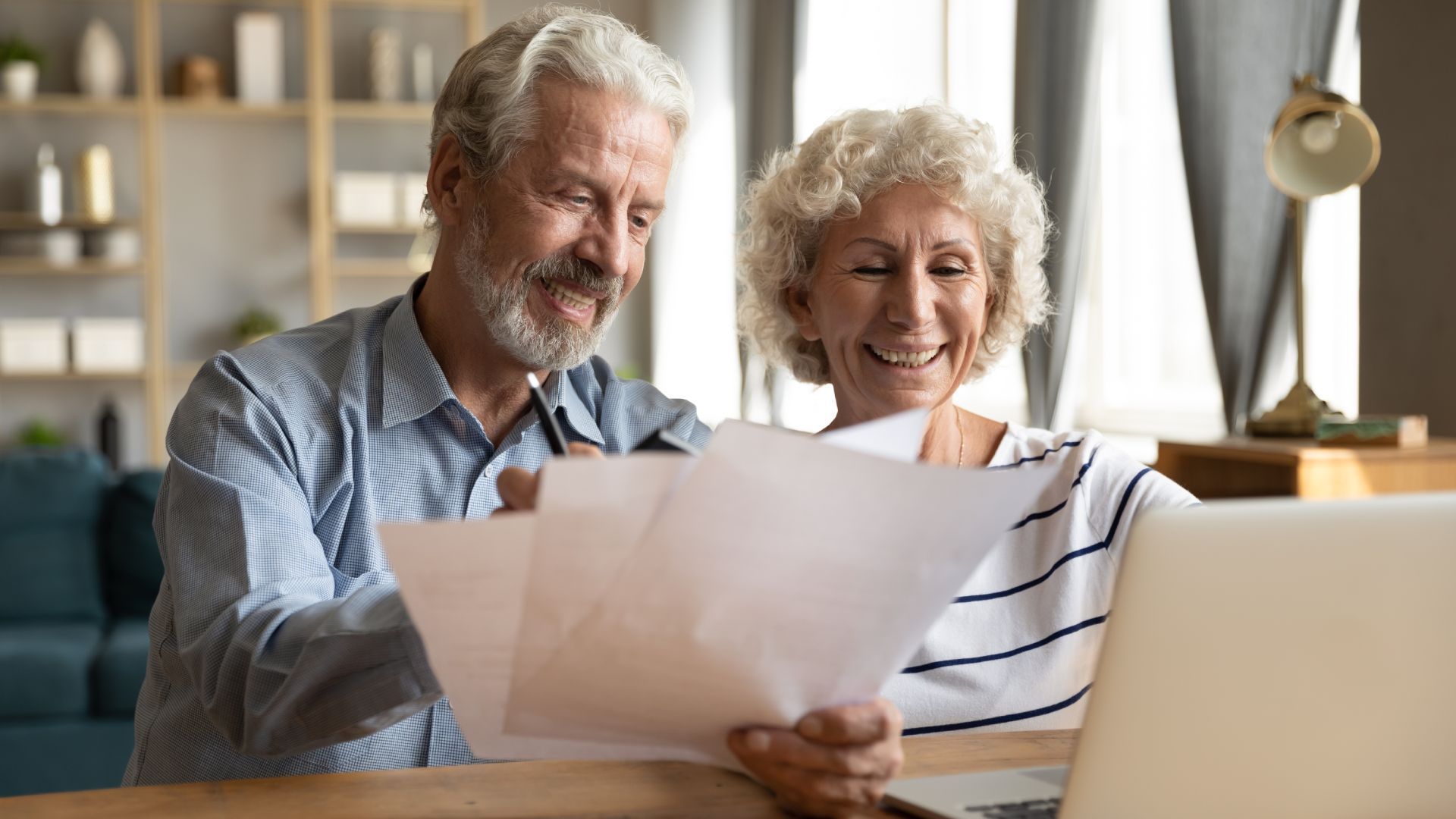 Older couple smiling, looking at papers with a laptop, in a home setting.
