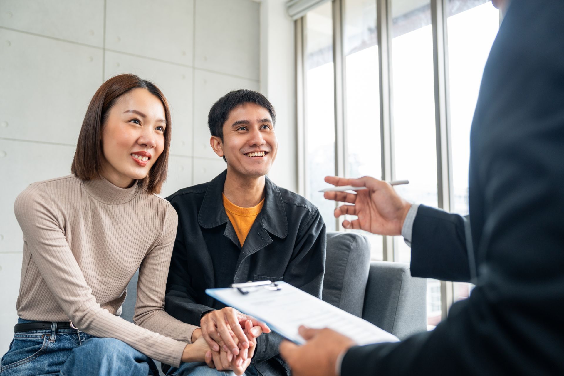 Couple smiles as they sit with a person holding a clipboard, indoors near a window.