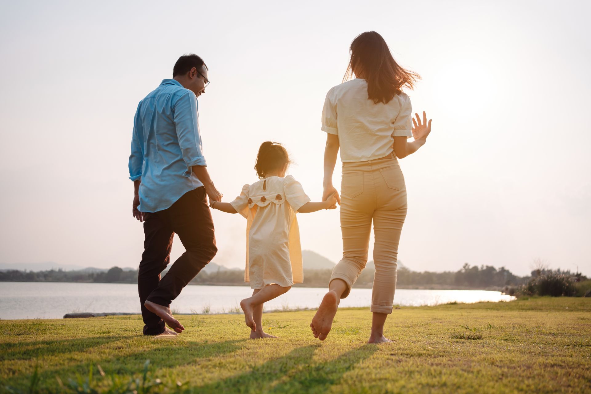 Family holding hands and walking on grass towards water, with sun in the background.