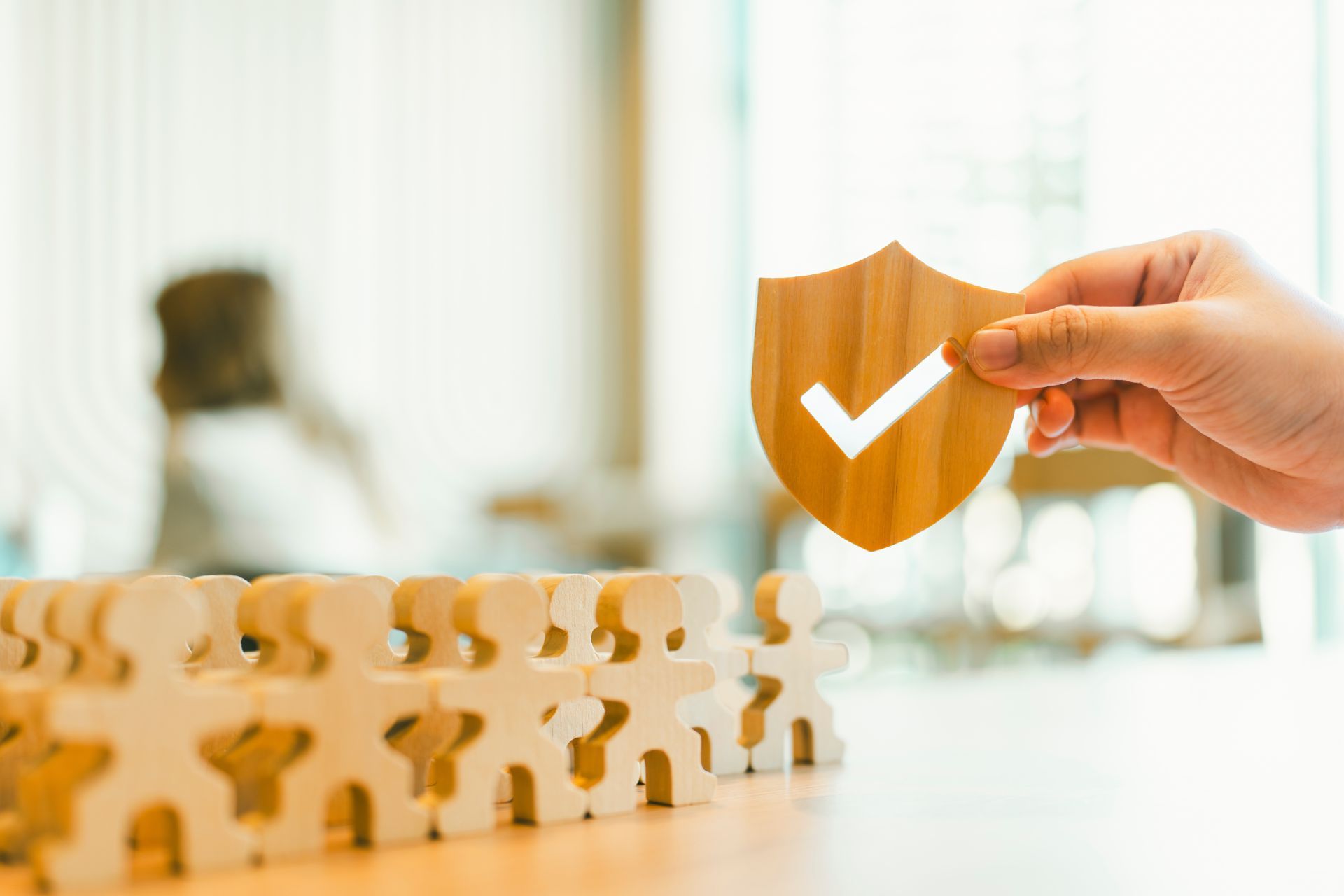 Hand holding a wooden shield with a checkmark above wooden figures, symbolizing protection or security.