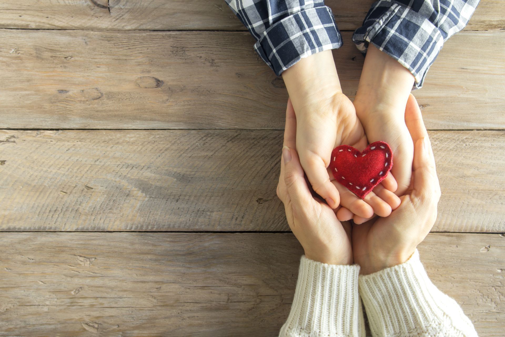 Hands holding a red heart on a wooden surface.