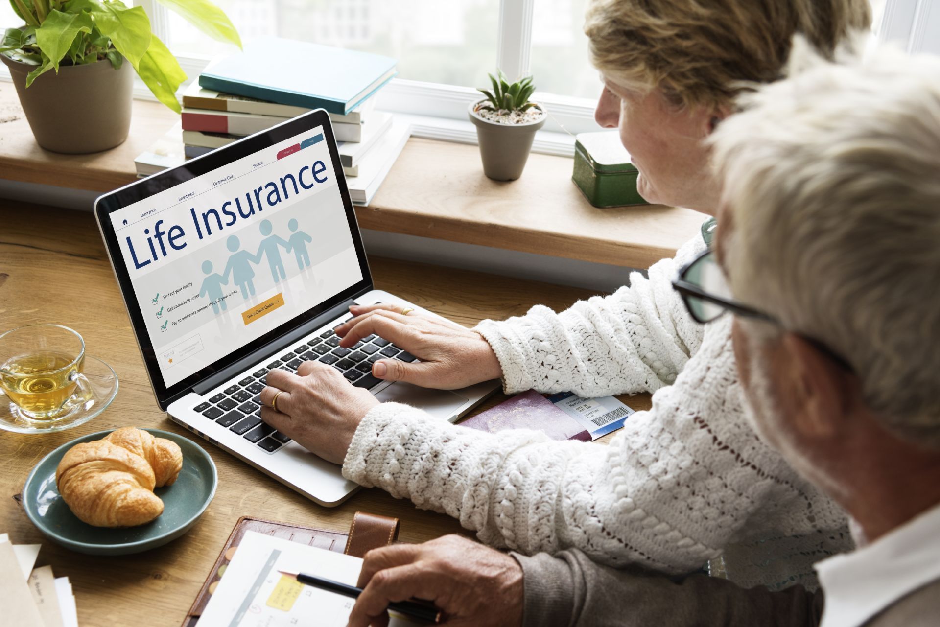 Couple using a laptop to research life insurance; croissant and tea visible.