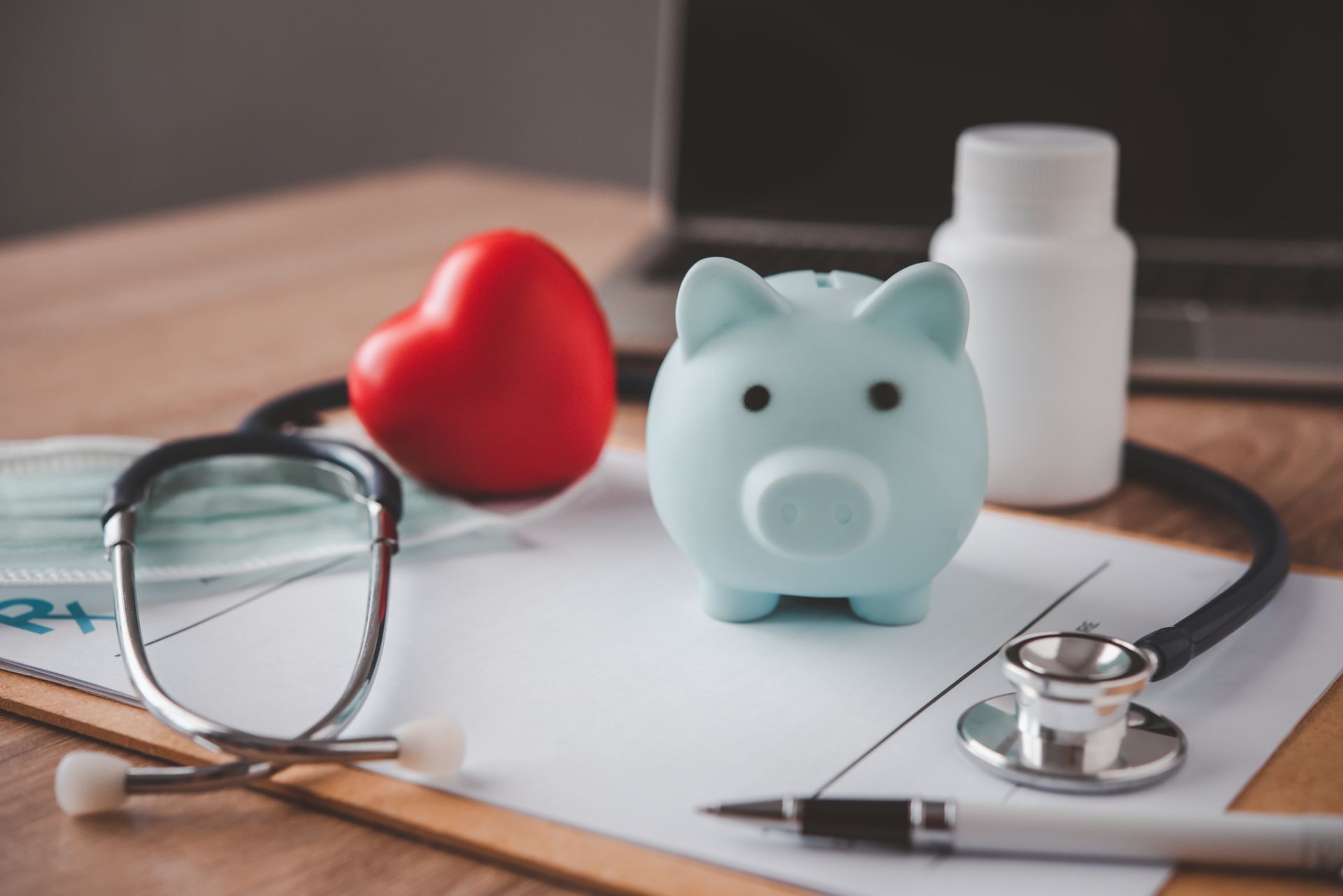 Piggy bank, stethoscope, red heart, and prescription on a desk, suggesting healthcare savings.