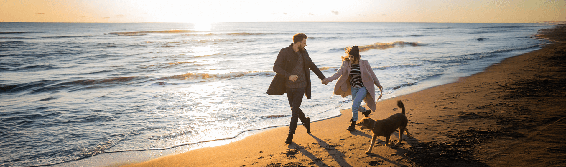 Couple holding hands, running on beach with dog. Sunlight on the water and sand.