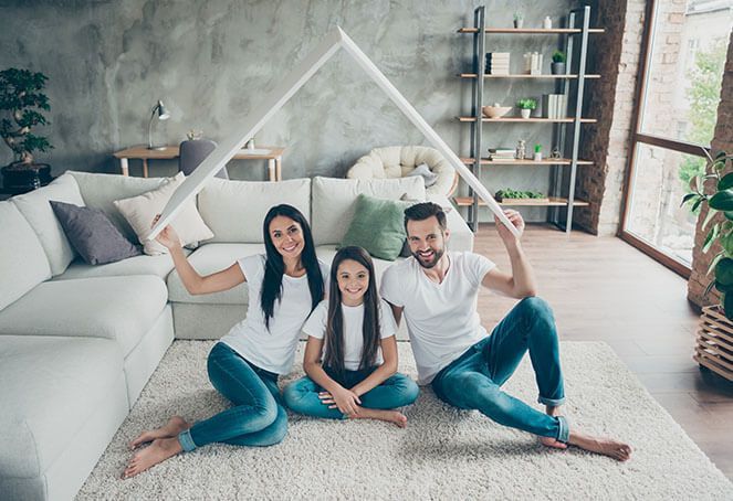 Family of three sitting on a rug under a roof-shaped cover in a living room; smiling.