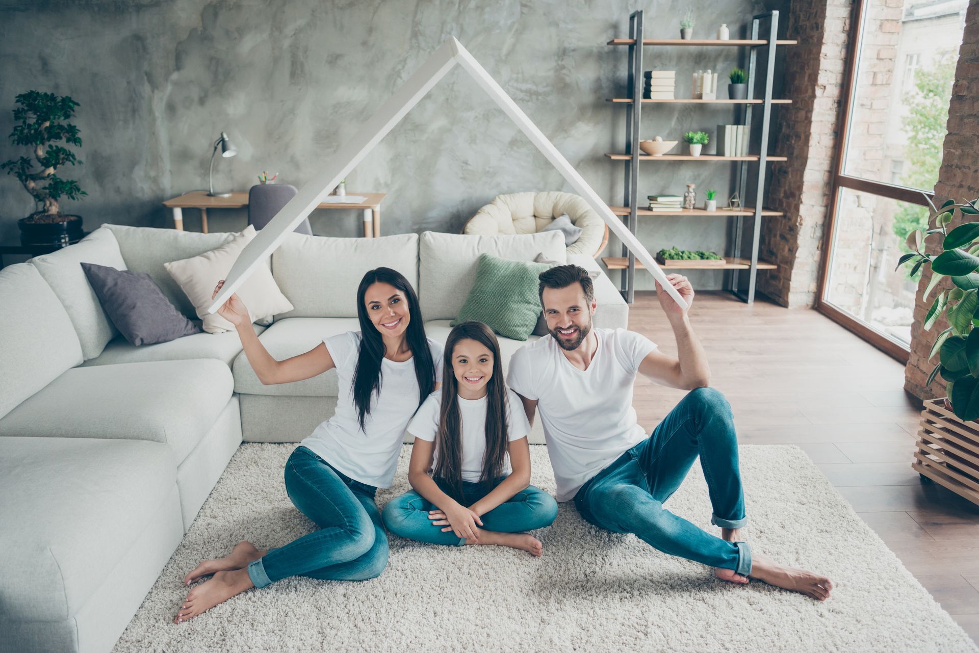 Family sitting on rug under a paper roof, smiling. Living room with couch and bookshelf.