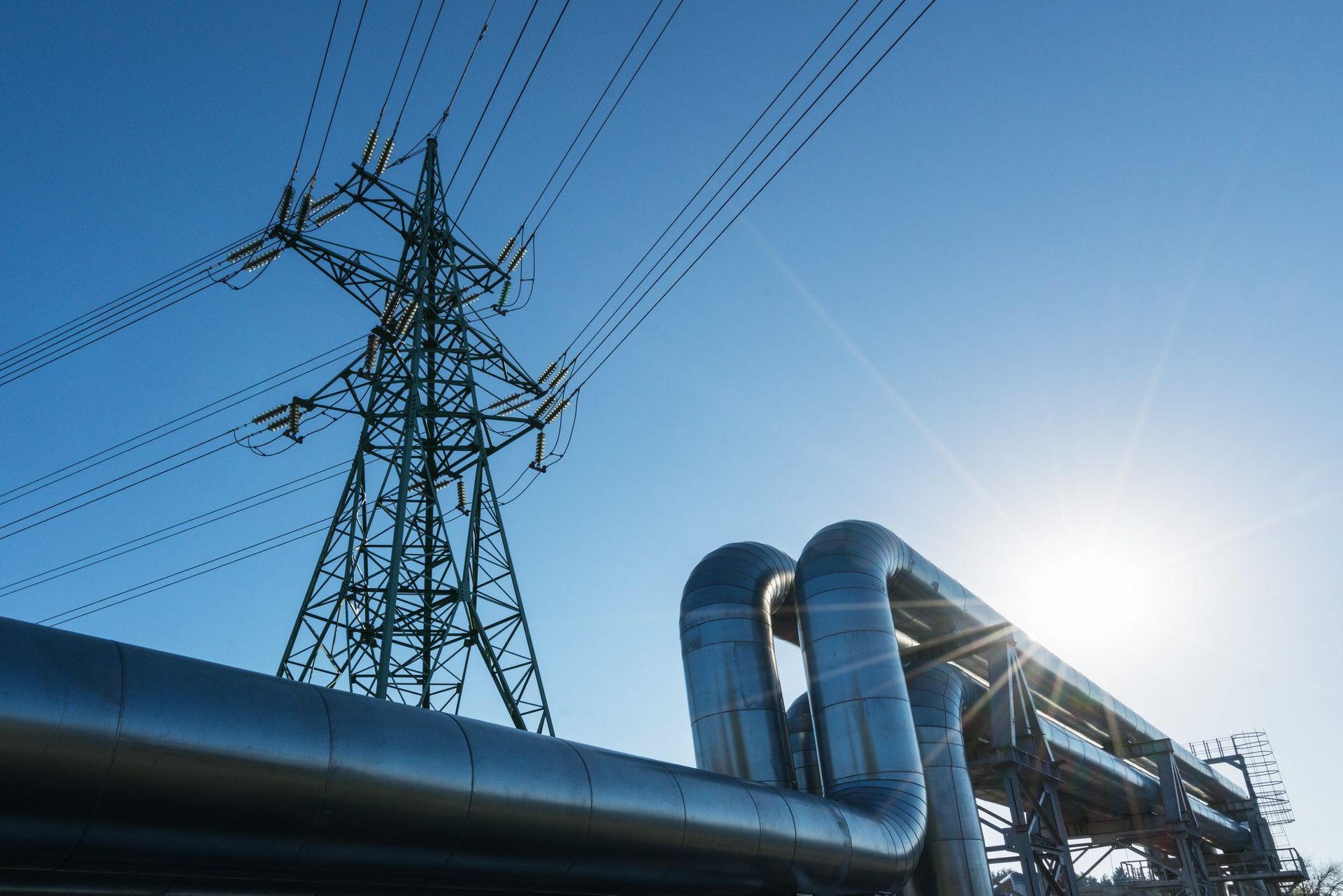Power lines and metal pipes against a bright blue sky, with the sun shining.