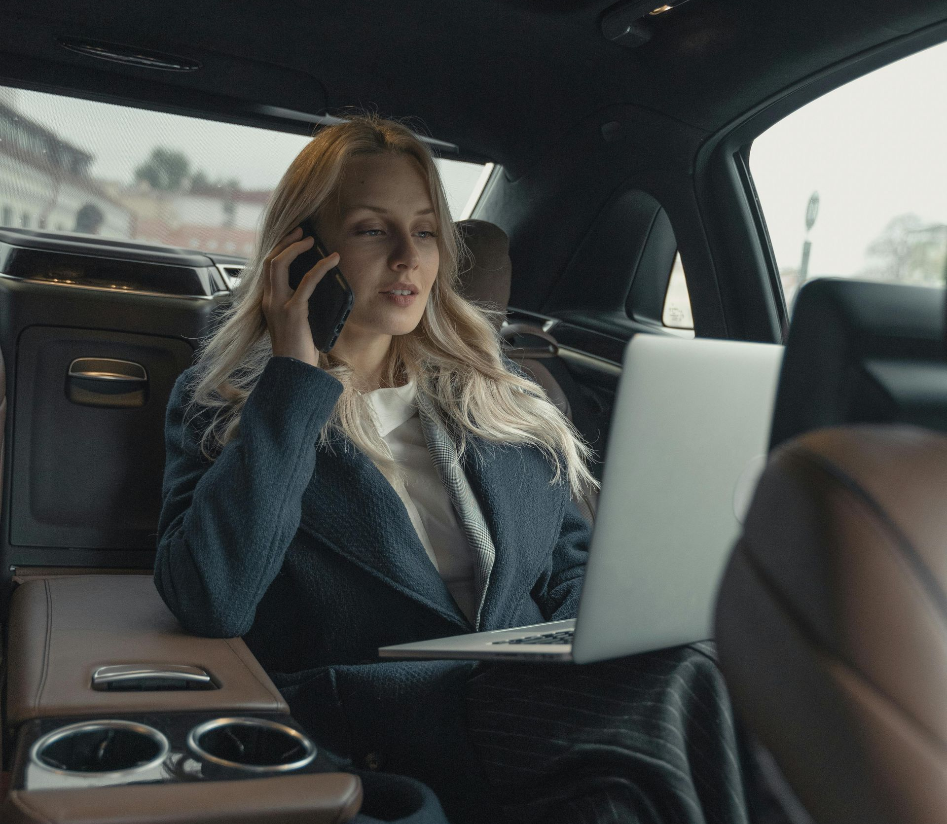 Woman in a car, talking on phone, working on a laptop. Interior shot.