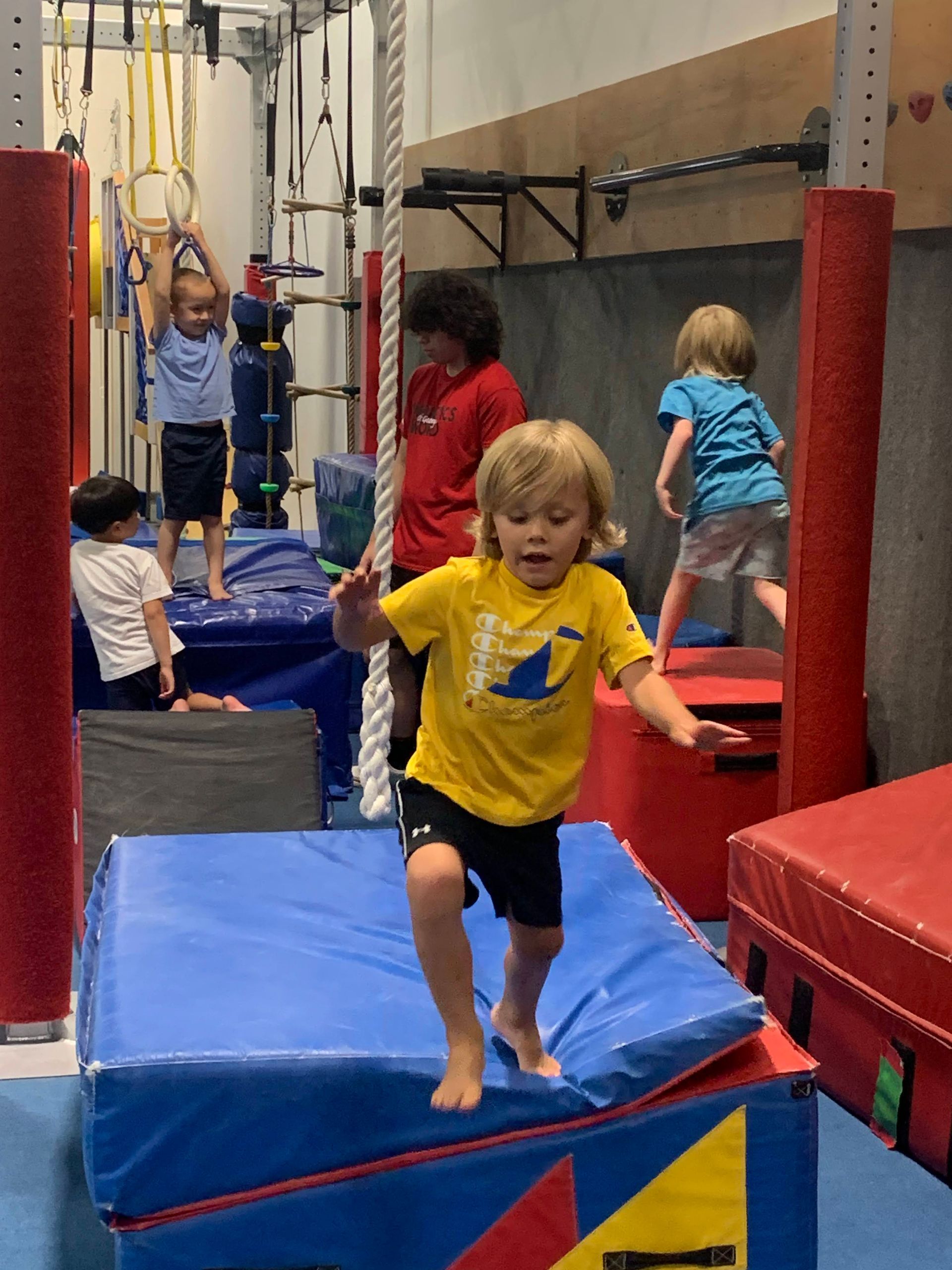 A young boy in a yellow shirt is walking on a blue mat in a gym.