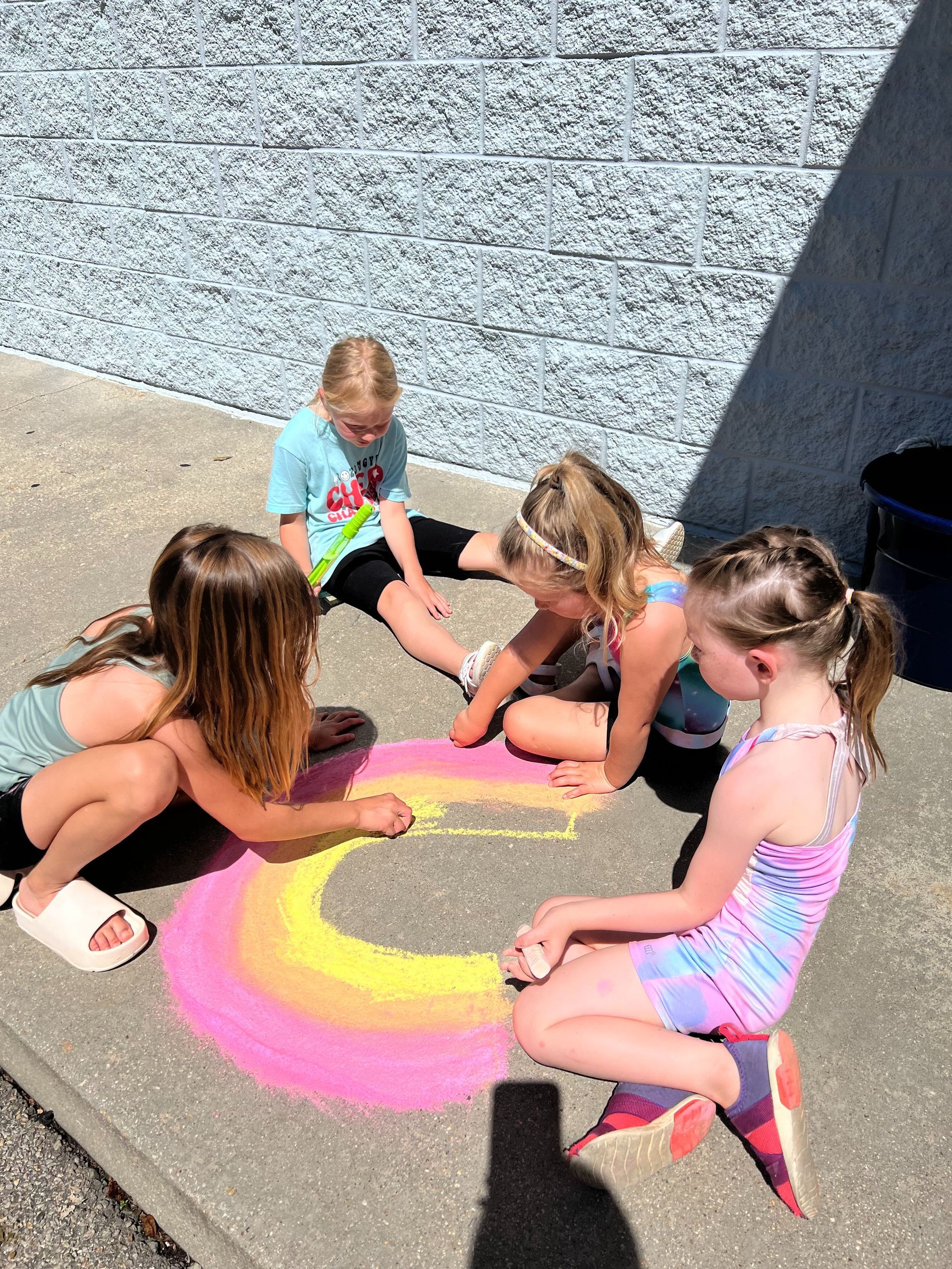 A group of young girls are sitting on the sidewalk drawing a rainbow with chalk.