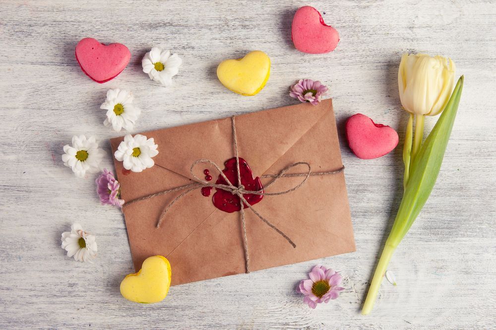 Brown envelope tied with twine, sealed with red wax, surrounded by heart-shaped macarons, flowers, and a yellow tulip on a white wooden surface.