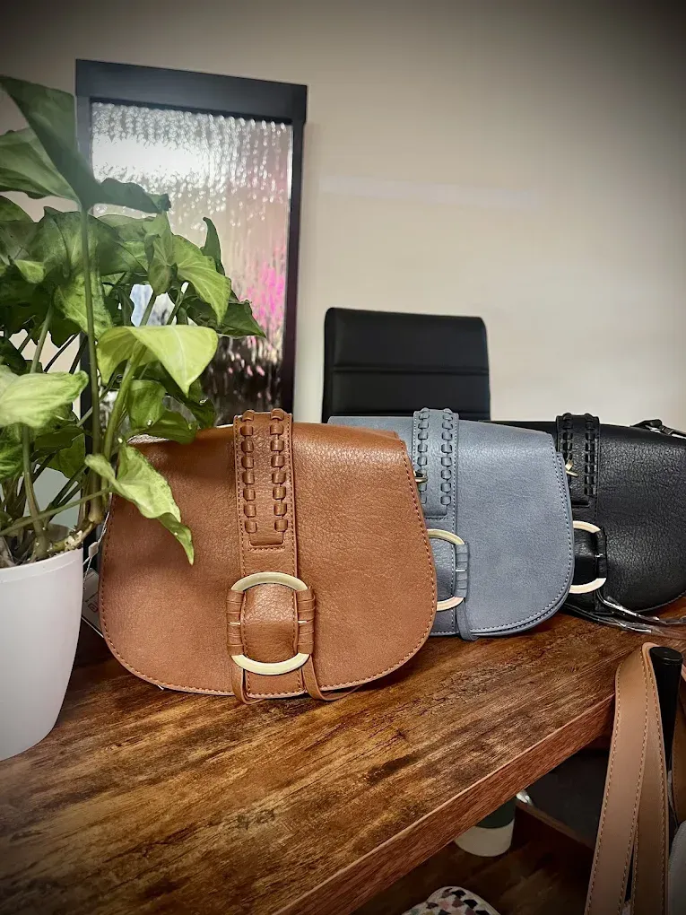 Three leather saddle bags in brown, blue, and black, displayed on a wooden surface with a plant and fountain backdrop.