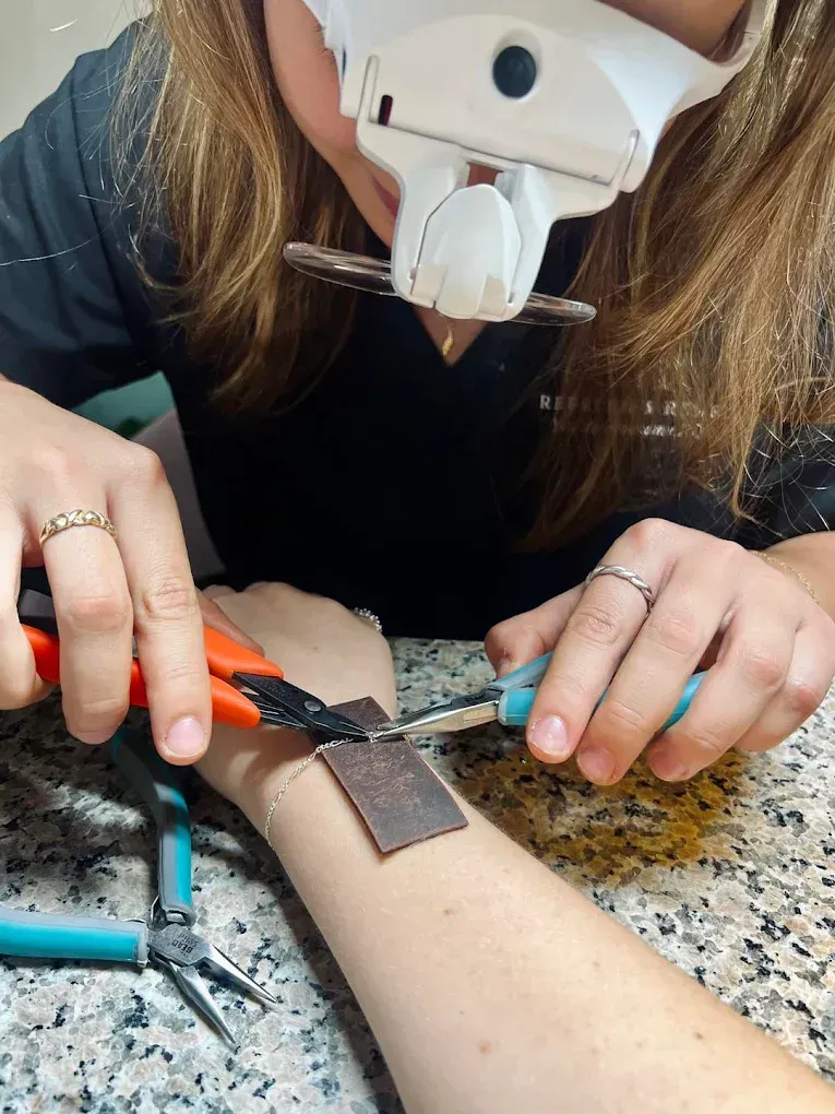Person using tools, with magnifying glasses, working on small metal squares attached to their wrist.