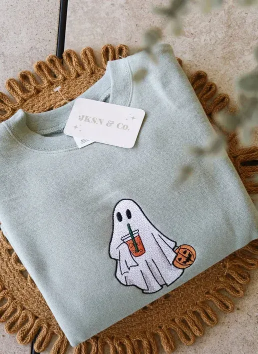 Green sweatshirt with ghost holding a drink and pumpkin, on a woven mat.