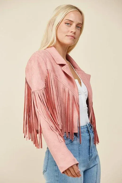 Woman in pink fringe jacket and blue jeans, standing against a beige background.