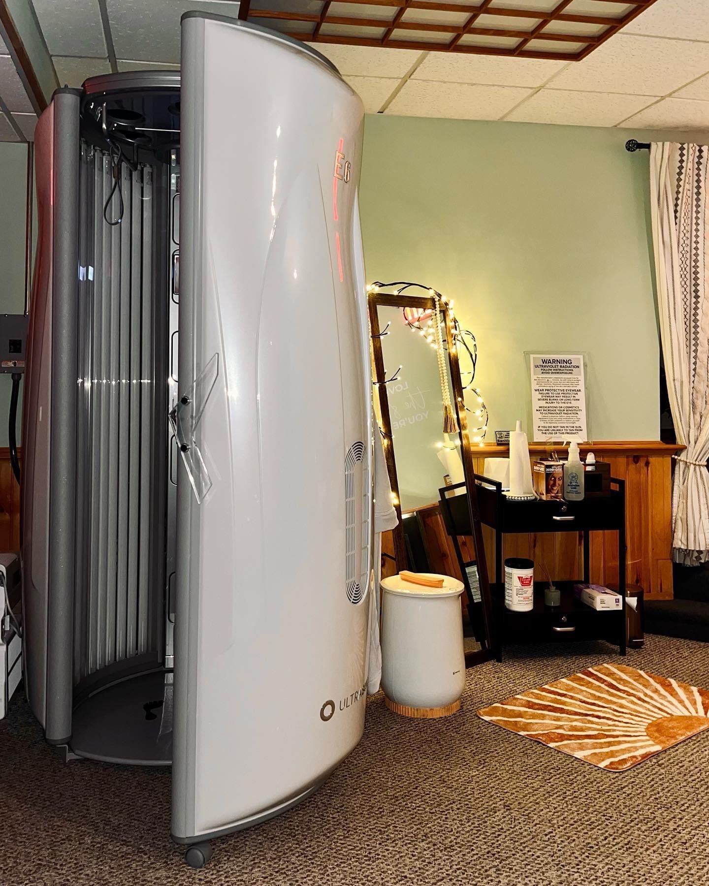 Tanning booth with open door in a room with a mirror, curtains, and small shelves, brown carpet, light green wall.