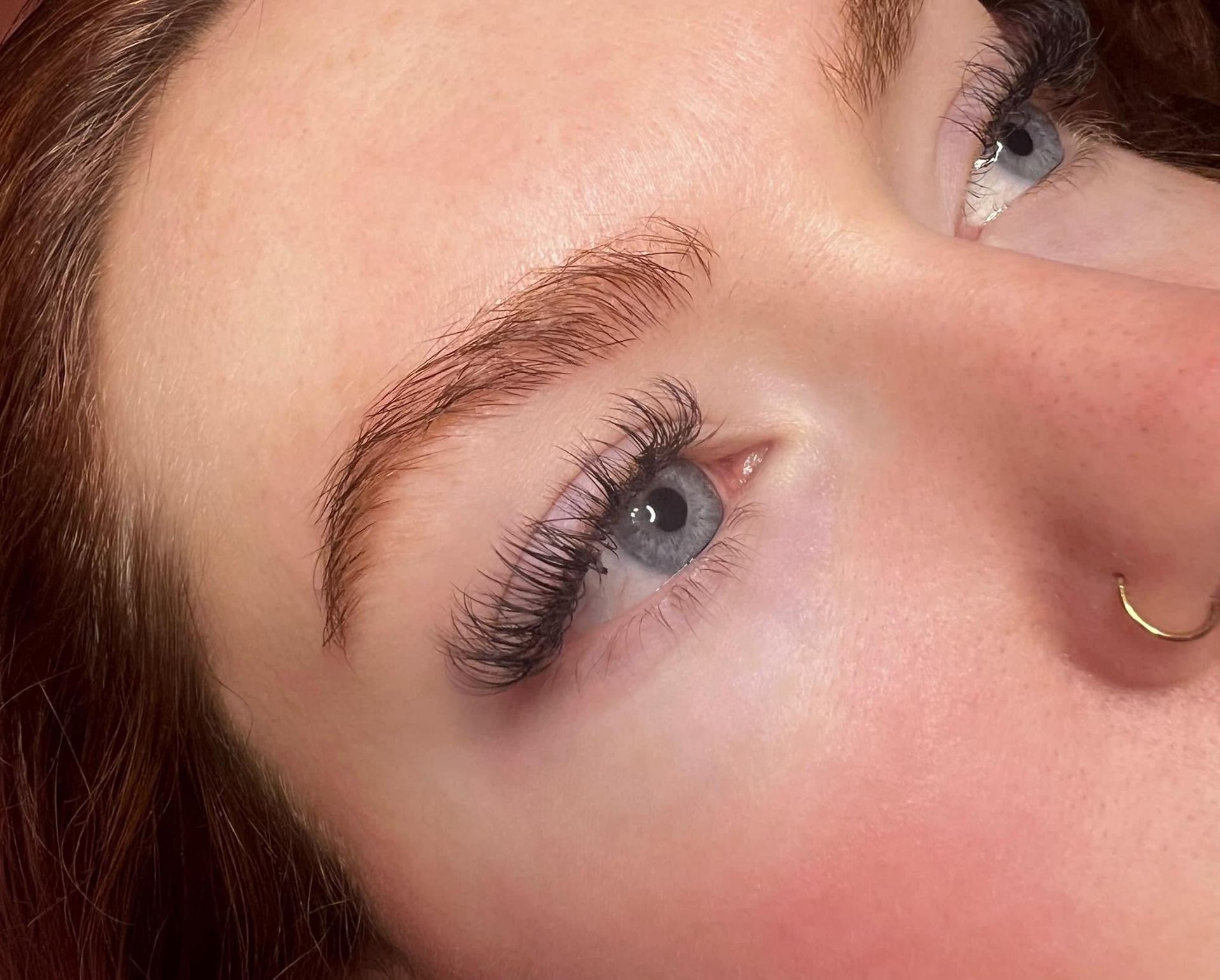 Close-up of a person's eye with eyebrow and eyelash enhancements, light skin, blue eye, gold nose ring.