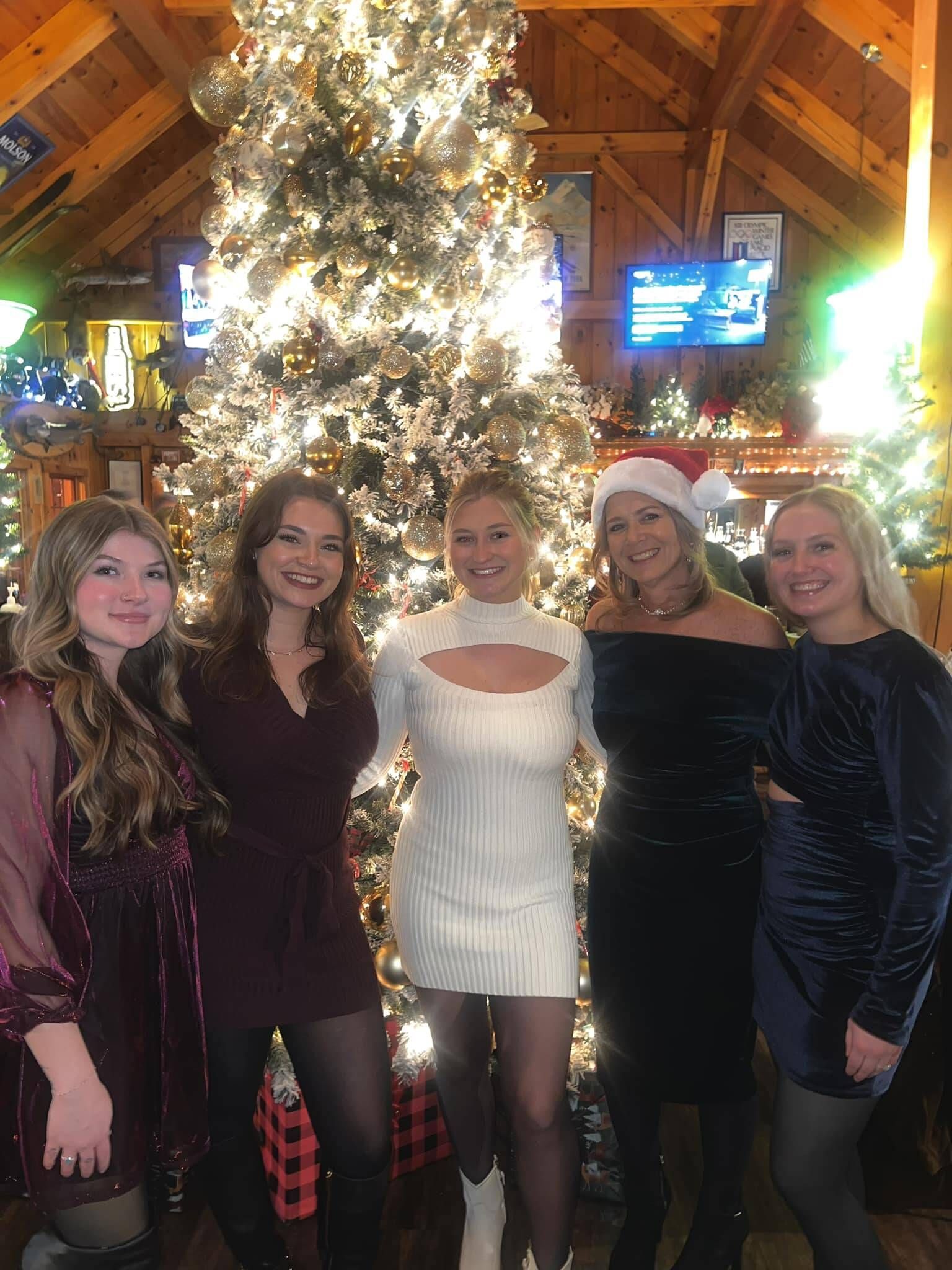 Five women smiling in front of a decorated Christmas tree. They wear dresses, tights, and a Santa hat. Wooden interior.