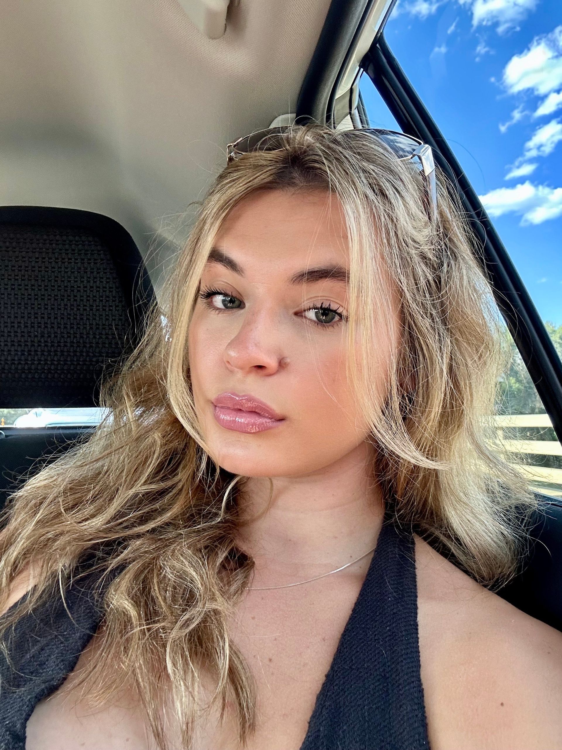 Woman in car, looking at the camera. Blonde hair, fair skin, black top, blue sky visible.