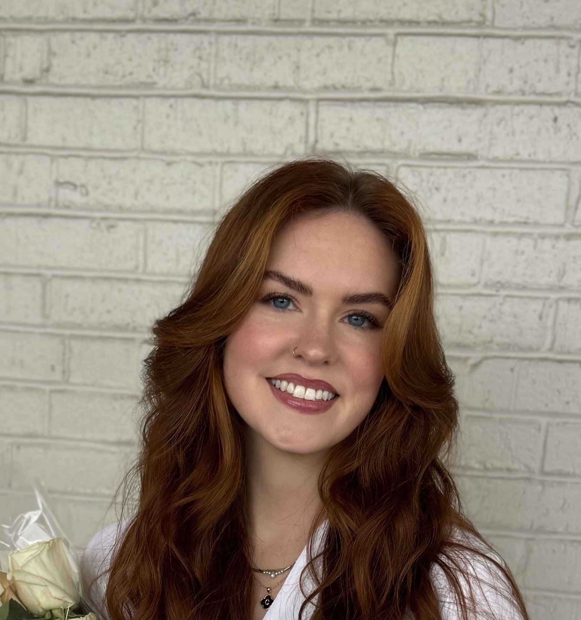 Woman with auburn hair smiles, holding flowers, in front of a white brick wall.