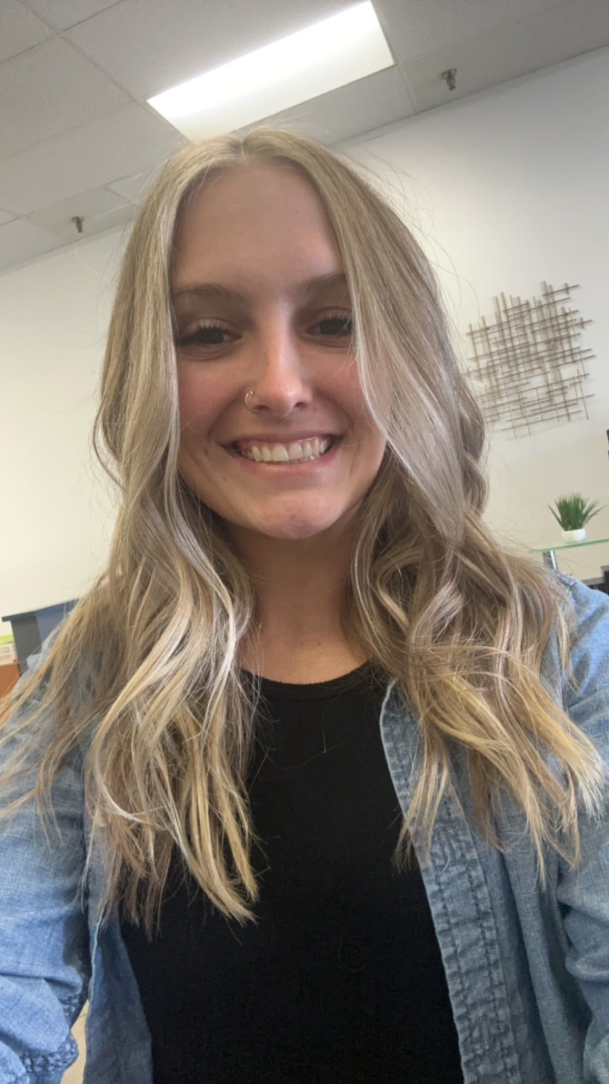 Woman with blonde wavy hair smiles at camera, wearing black shirt and denim jacket.